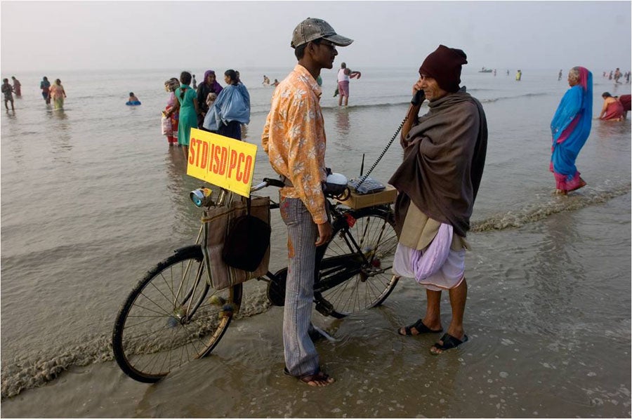 A man makes a phone call from a bicycle while standing in the ocean.