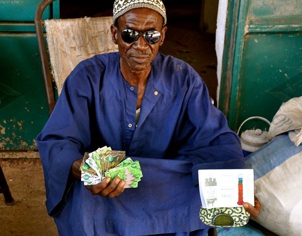 An elderly Senegalese farmer proudly displays the saving 'voucher' cards he has brought from myAgro over many months, and about to finish his savings milestone (displayed on thermometer!)