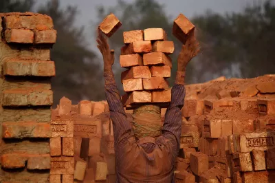 A brick worker carries bricks in a stack.