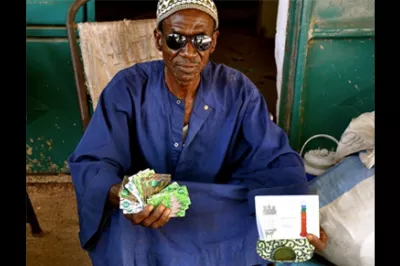 An elderly Senegalese farmer proudly displays the saving 'voucher' cards he has brought from myAgro over many months, and about to finish his savings milestone (displayed on thermometer!)