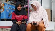 two women with headscarves sit on the curb outside a shop while looking down at their mobile phones